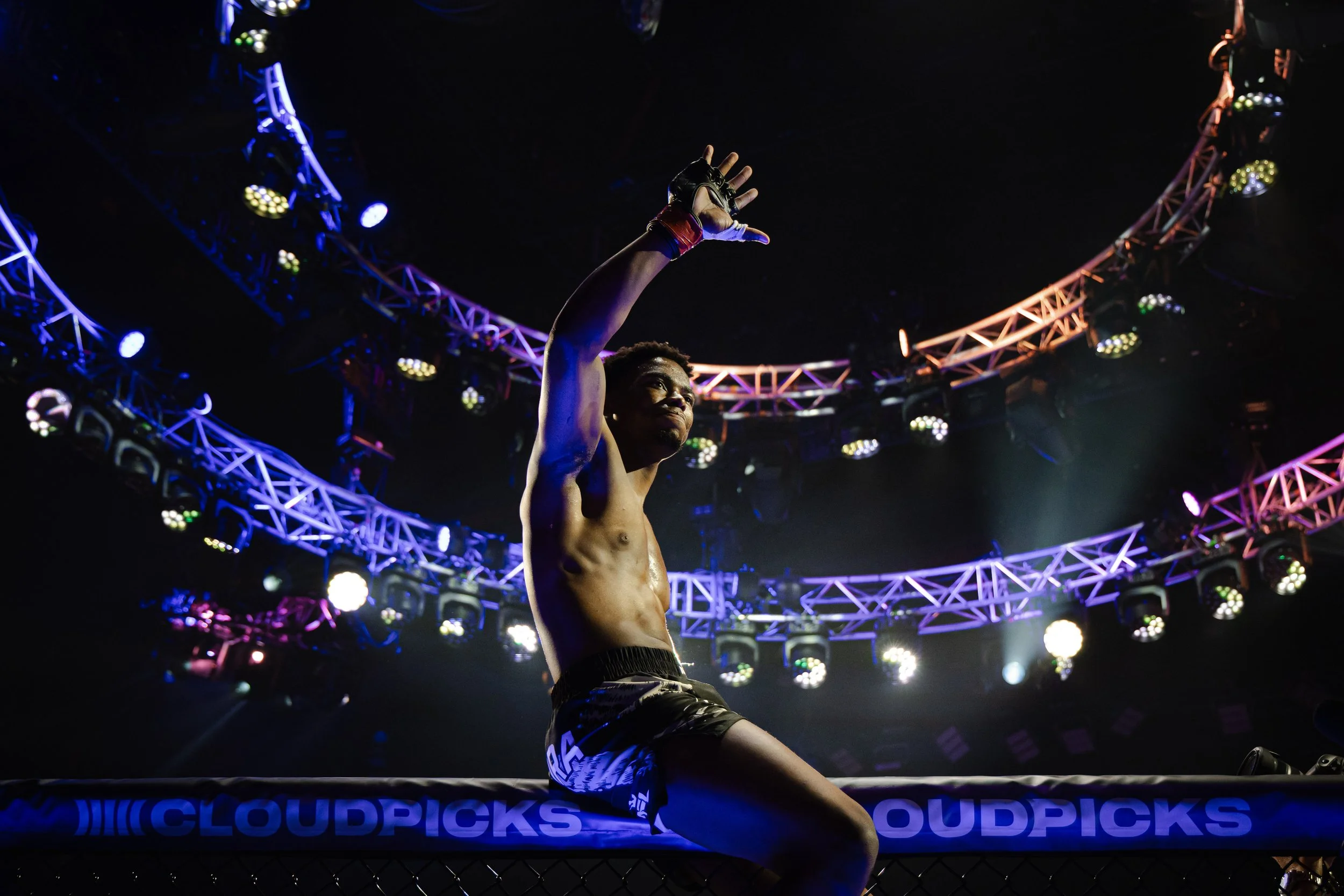 Een mannelijke vechter in een ring,  bij een mixed martial arts evenement, met een indrukwekkende houding en een opgeheven hand, omringd door kleurrijk verlichte arceringen en decoraties.
