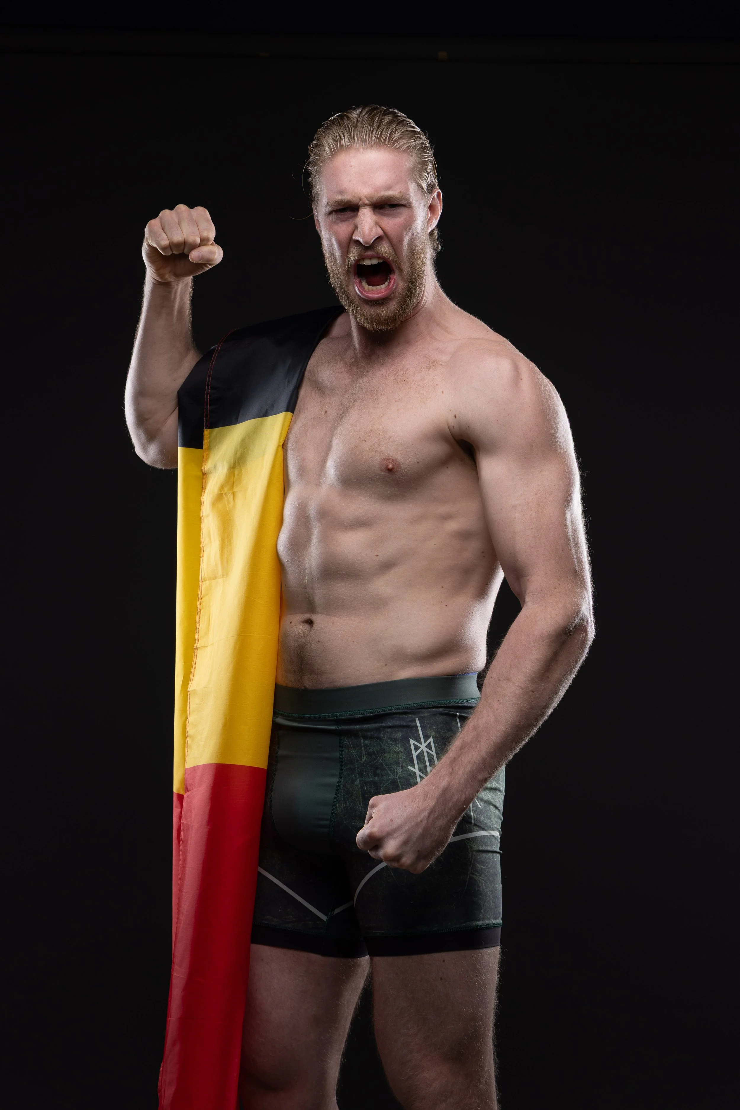 Een man met een behaarde borst en korte broek die een Belgische vlag en een Rode Duivels-jasje draagt, met zijn vuist gebald en schreeuwend.