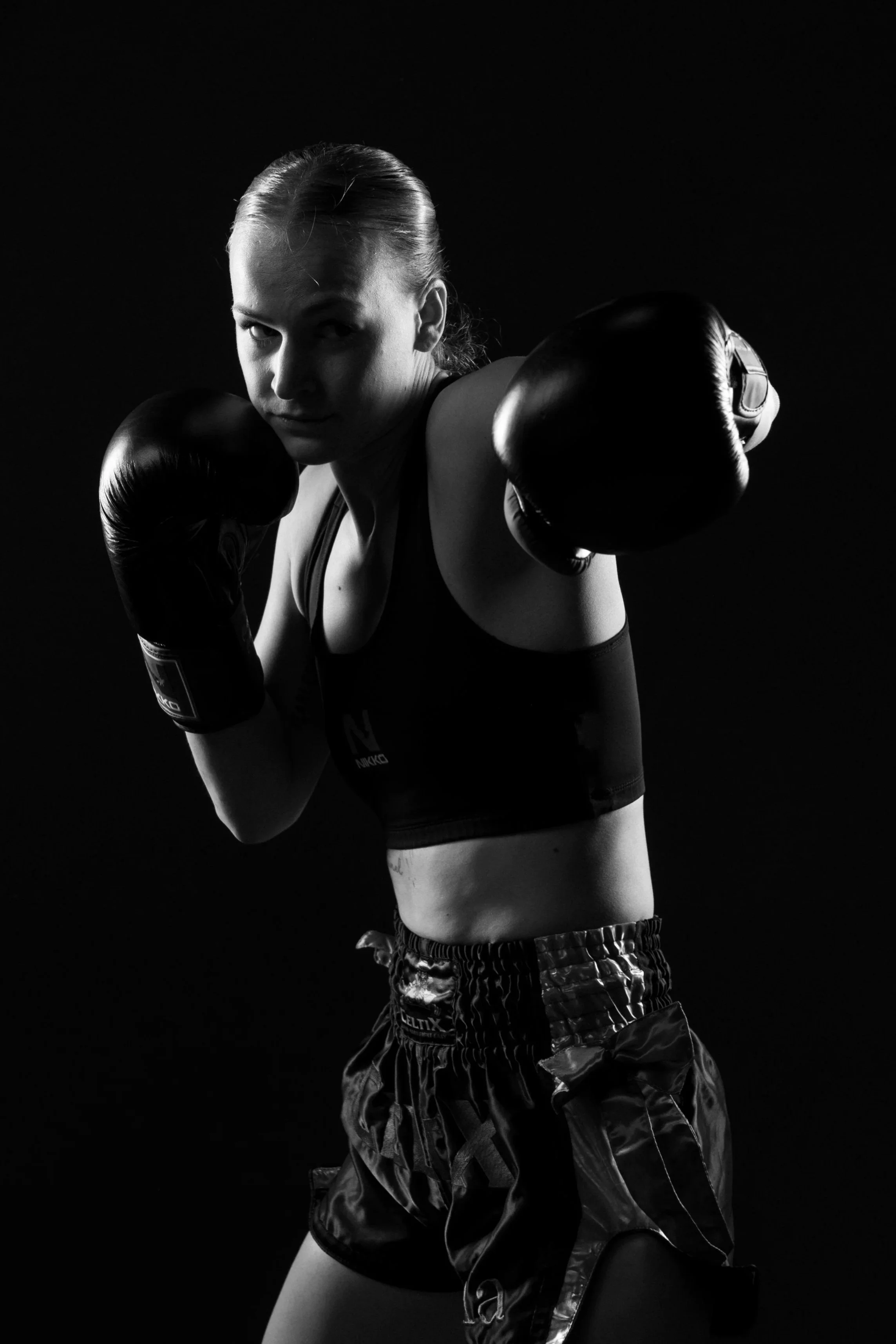 Zwarte en witte foto van een vrouwelijke bokser in een pose met haar handen omhoog, gedragen in bokshandschoenen, sportkleding en trainingsshorts, met een donkere achtergrond.