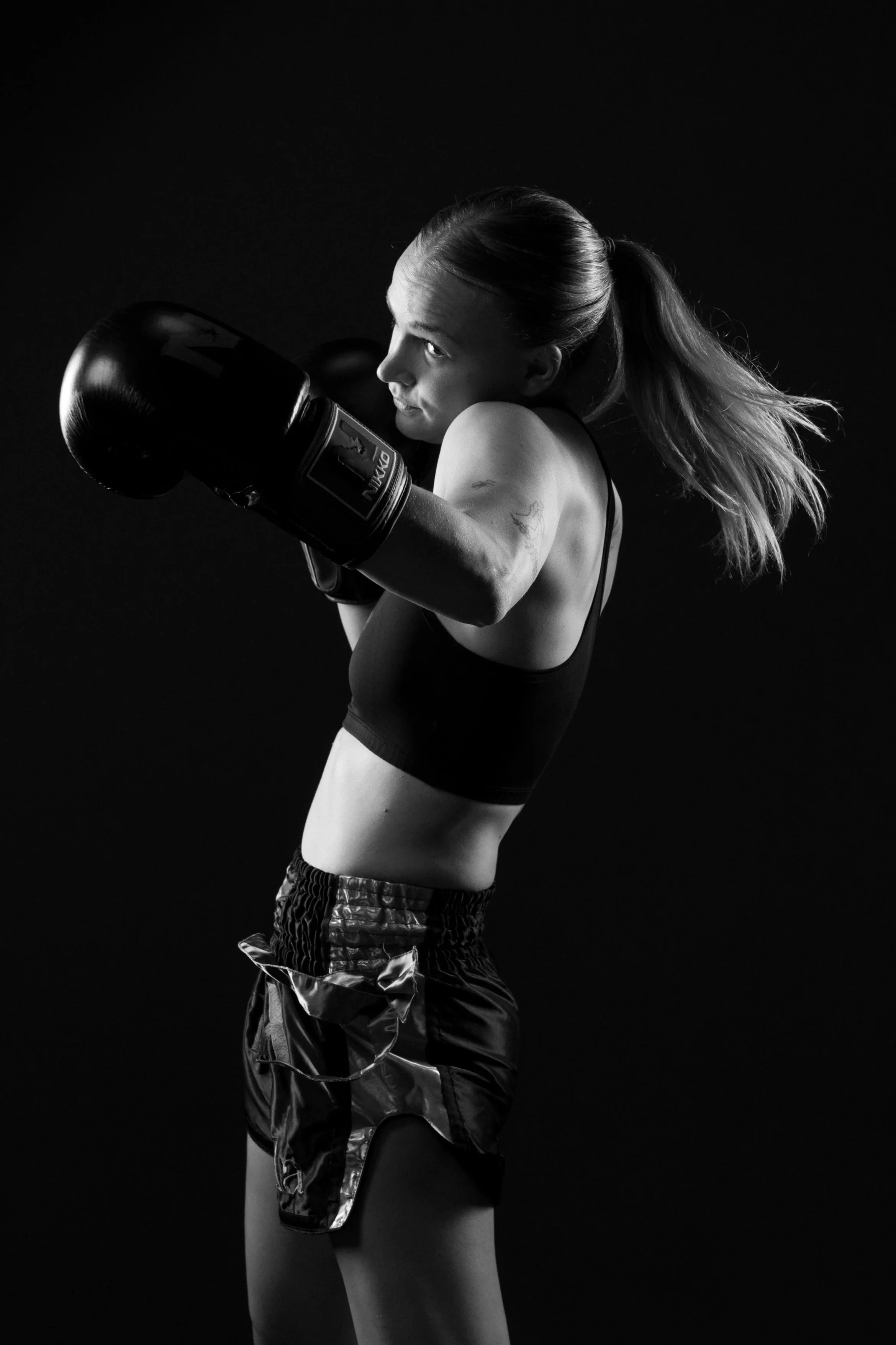 Zwart-wit foto van een vrouw in een sportoutfit met bokshandschoenen in een sportschoolomgeving.