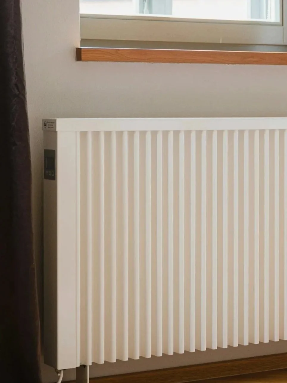 White electrical storage heater with vertical fins mounted on a wall below a window with a wooden window ledge.