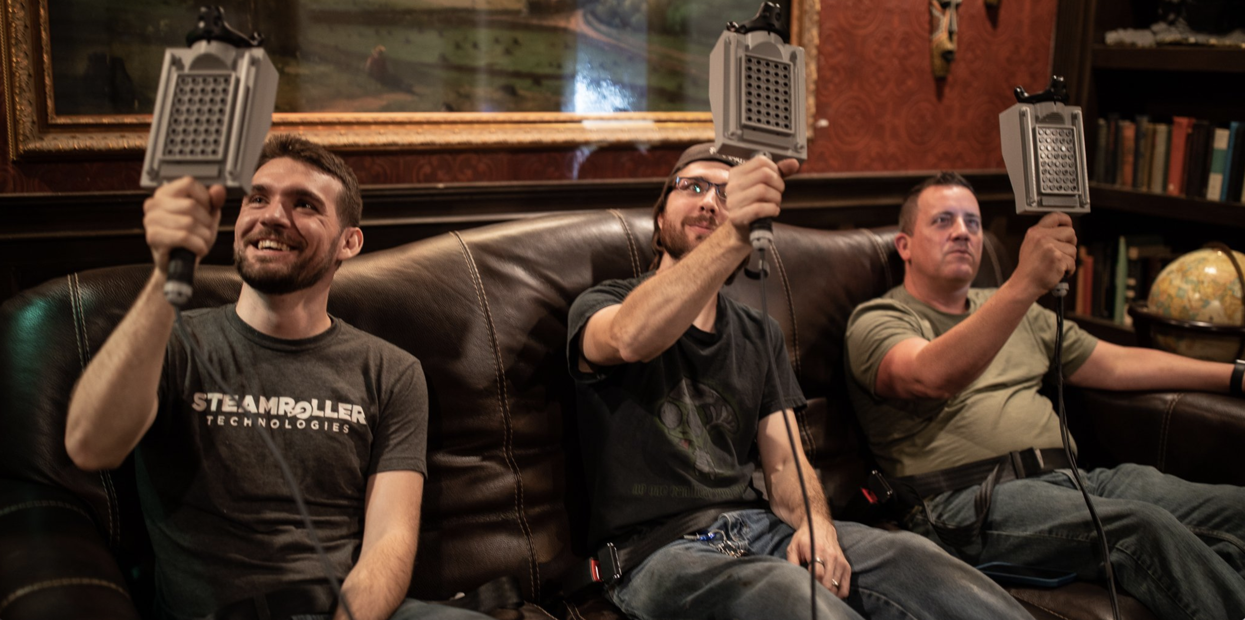 Three men sitting on a brown couch motion platform with handheld digital devices held up