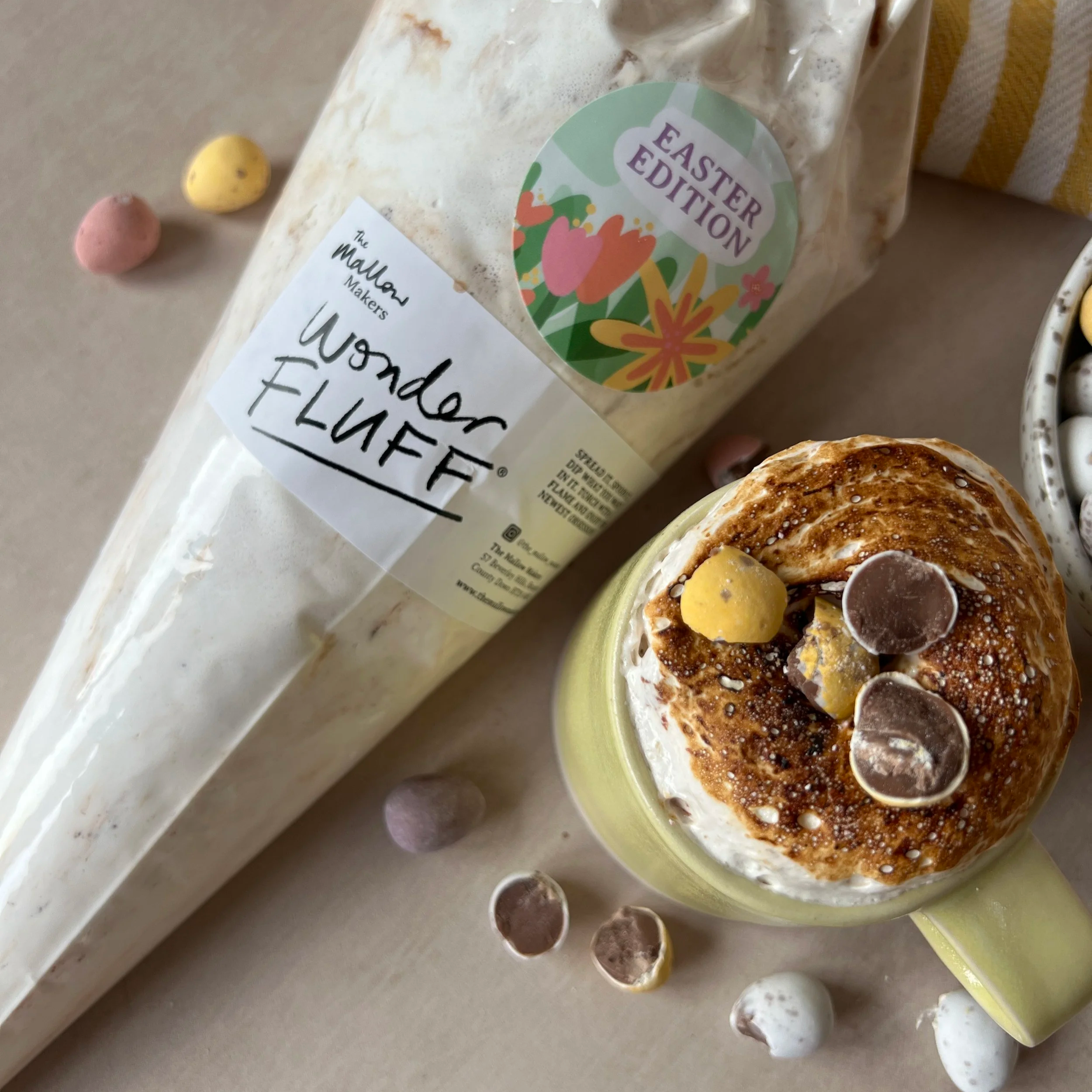 Looking+down+on+a+mug+of+hot+chocolate+topped+with+Mini+Eggs+Wonder+Fluff+and+crushed+Mini+Eggs+with+a+piping+bag+of+Wonder+Fluff+on+a+striped+yellow+table+cloth.png