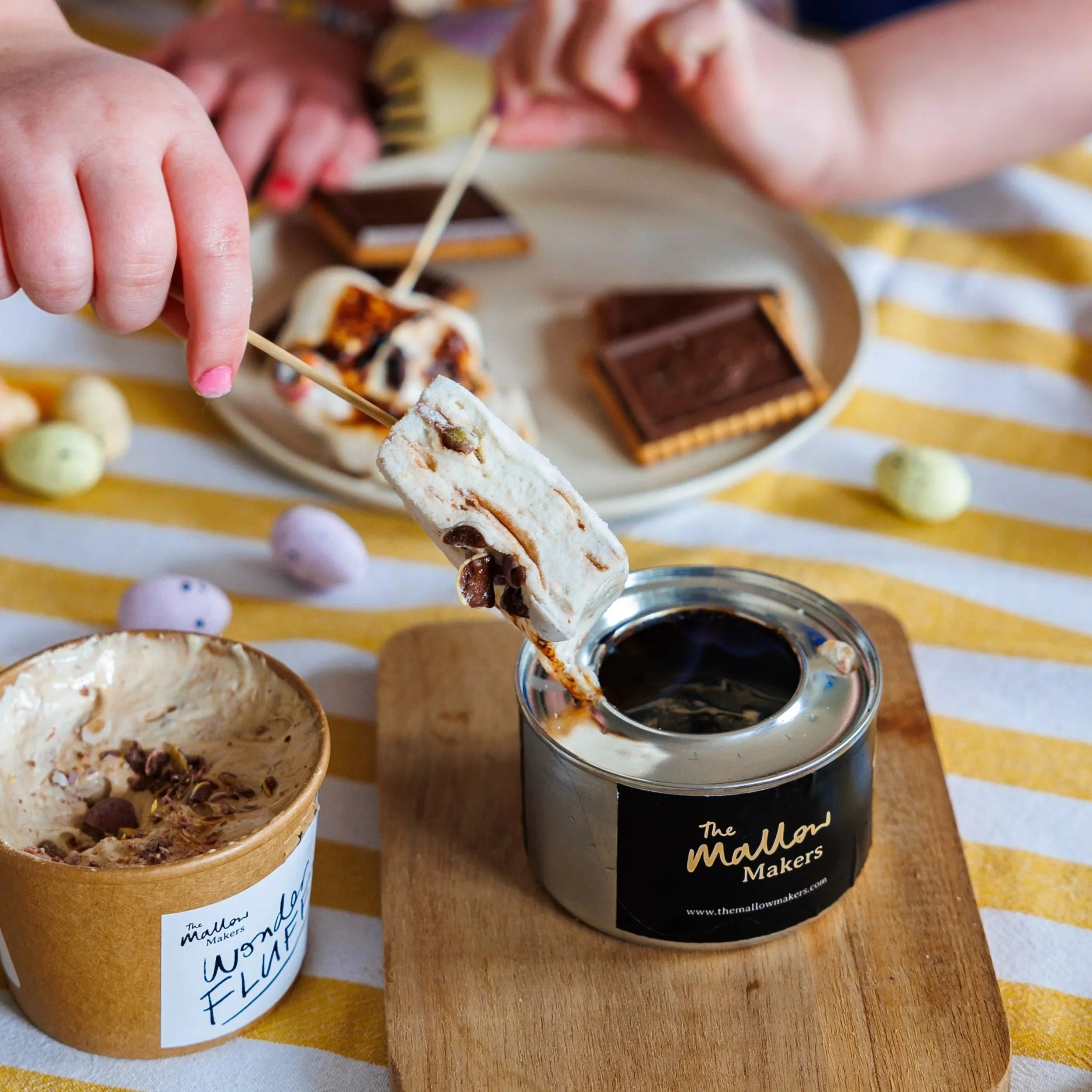 children+making+smores+over+a+The+Mallow+Makers+mini+gel+burner+on+a+wooden+chopping+board+on+a+striped+yellow+table+cloth+beside+a+tub+of+mini+eggs+wonder+fluff.jpg