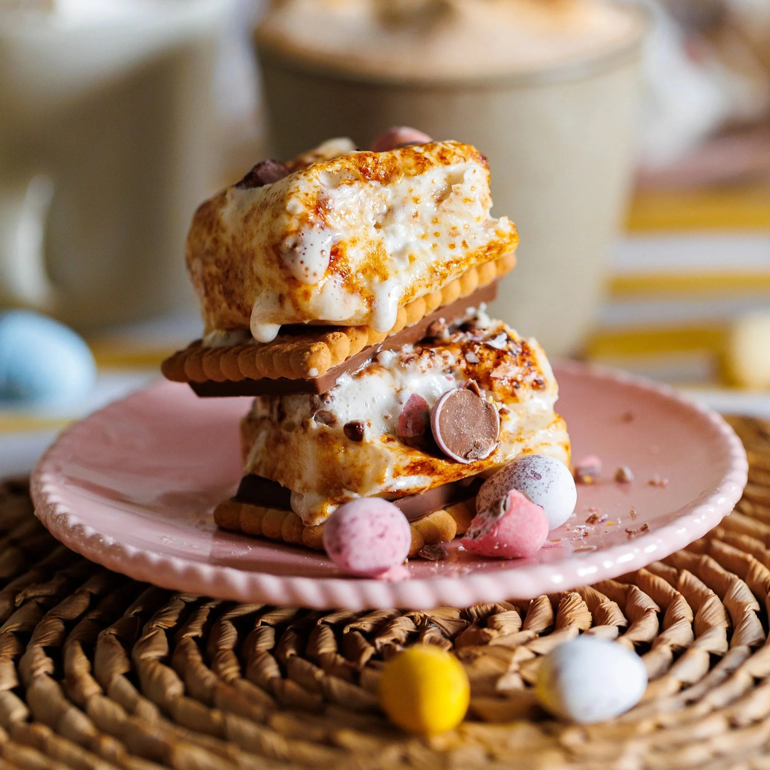 Toasted+marshmallows+and+mini+eggs+stacked+between+biscuits+on+top+of+a+pink+plate+on+a+wicker+place+mat.jpg