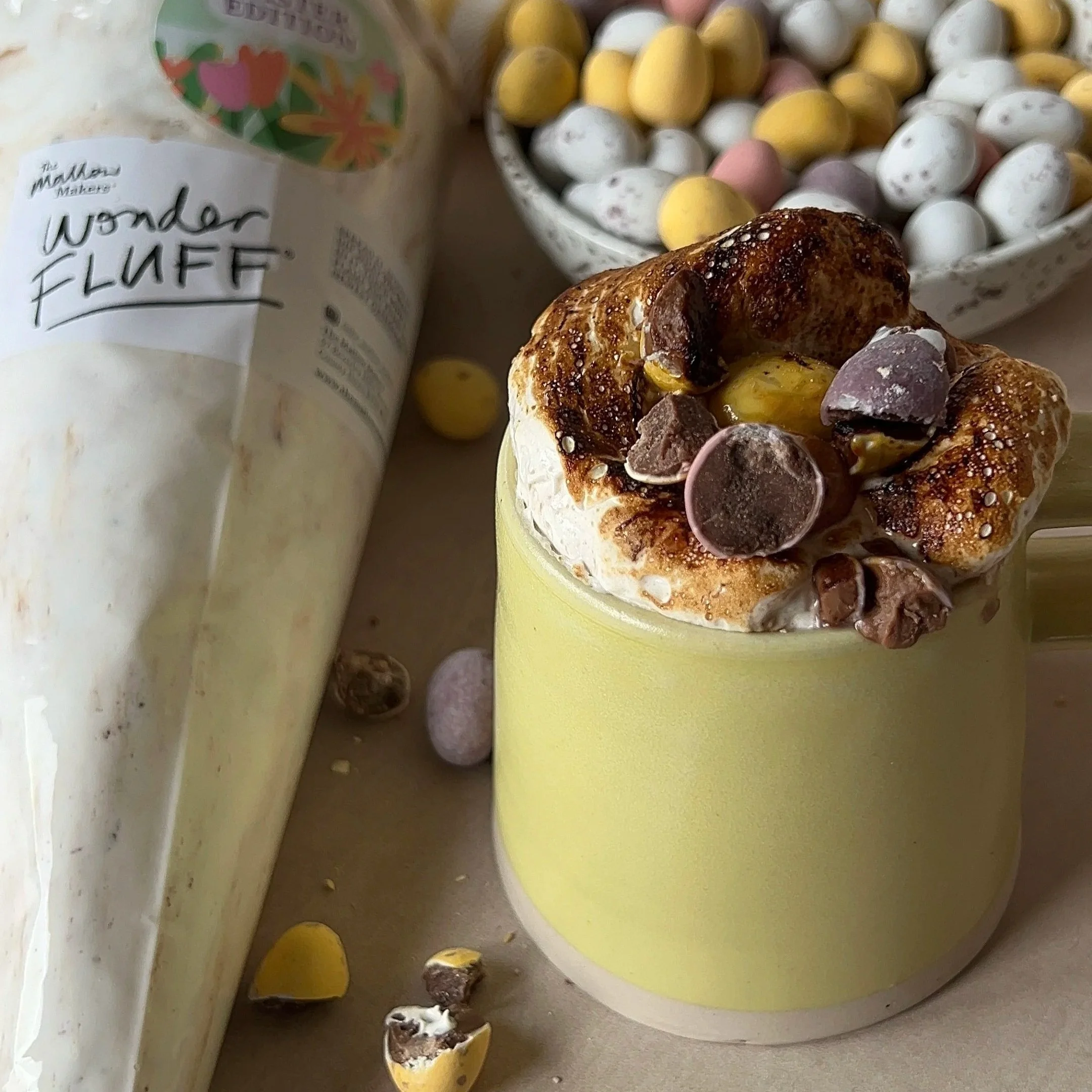 A+mug+of+hot+chocolate+topped+with+Mini+Eggs+Wonder+Fluff+and+crushed+Mini+Eggs+with+a+piping+bag+of+Wonder+Fluff+on+a+striped+yellow+table+cloth.jpg