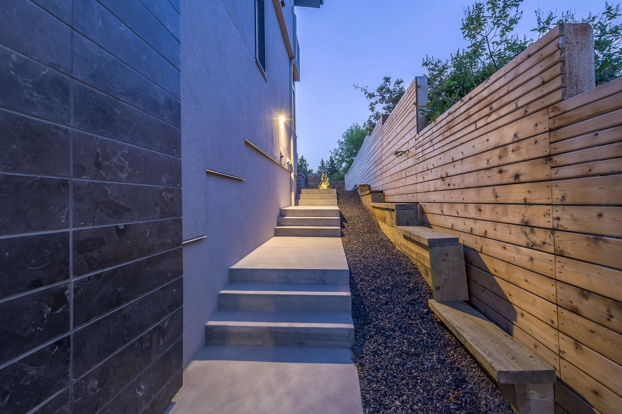 Side yard with concrete stairs, wooden retaining wall, green foliage, and exterior house wall with a light