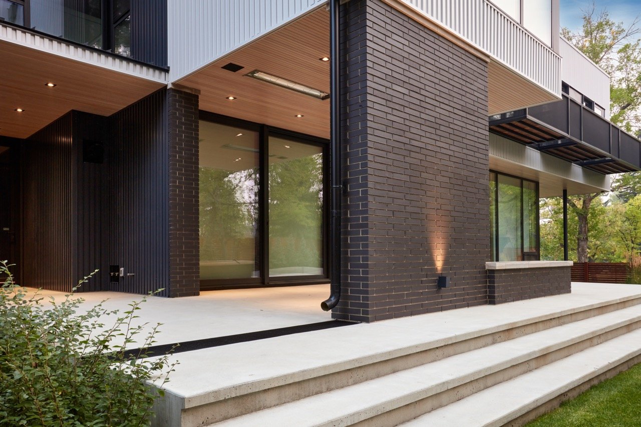 Modern house exterior with black brick and wood siding, large sliding glass doors, and a concrete patio with steps surrounded by greenery.