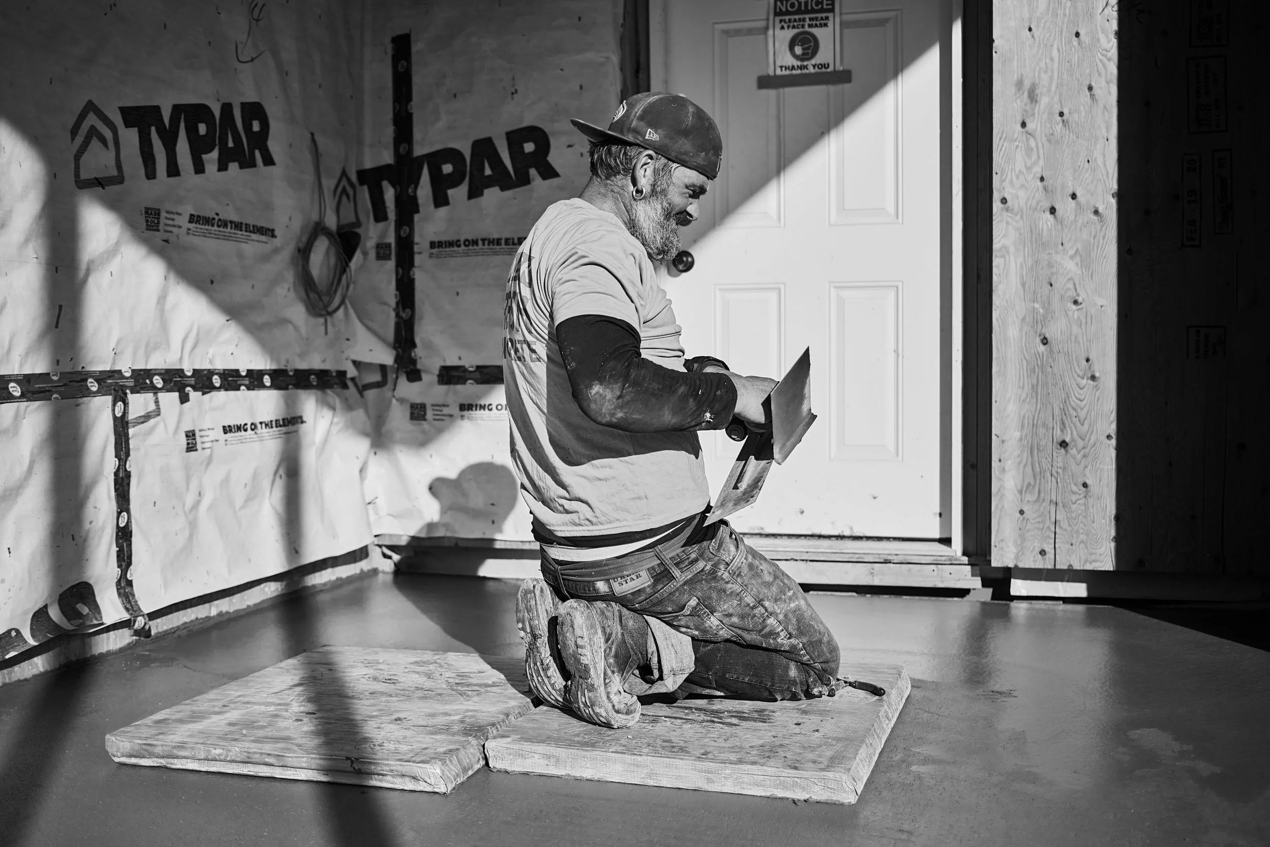 A man with a beard, earrings, and a cap kneels on a wooden platform while working on a construction project inside a building. The background shows insulation material with the brand name 'TYPAR' and a door with a notice to wear a face mask.