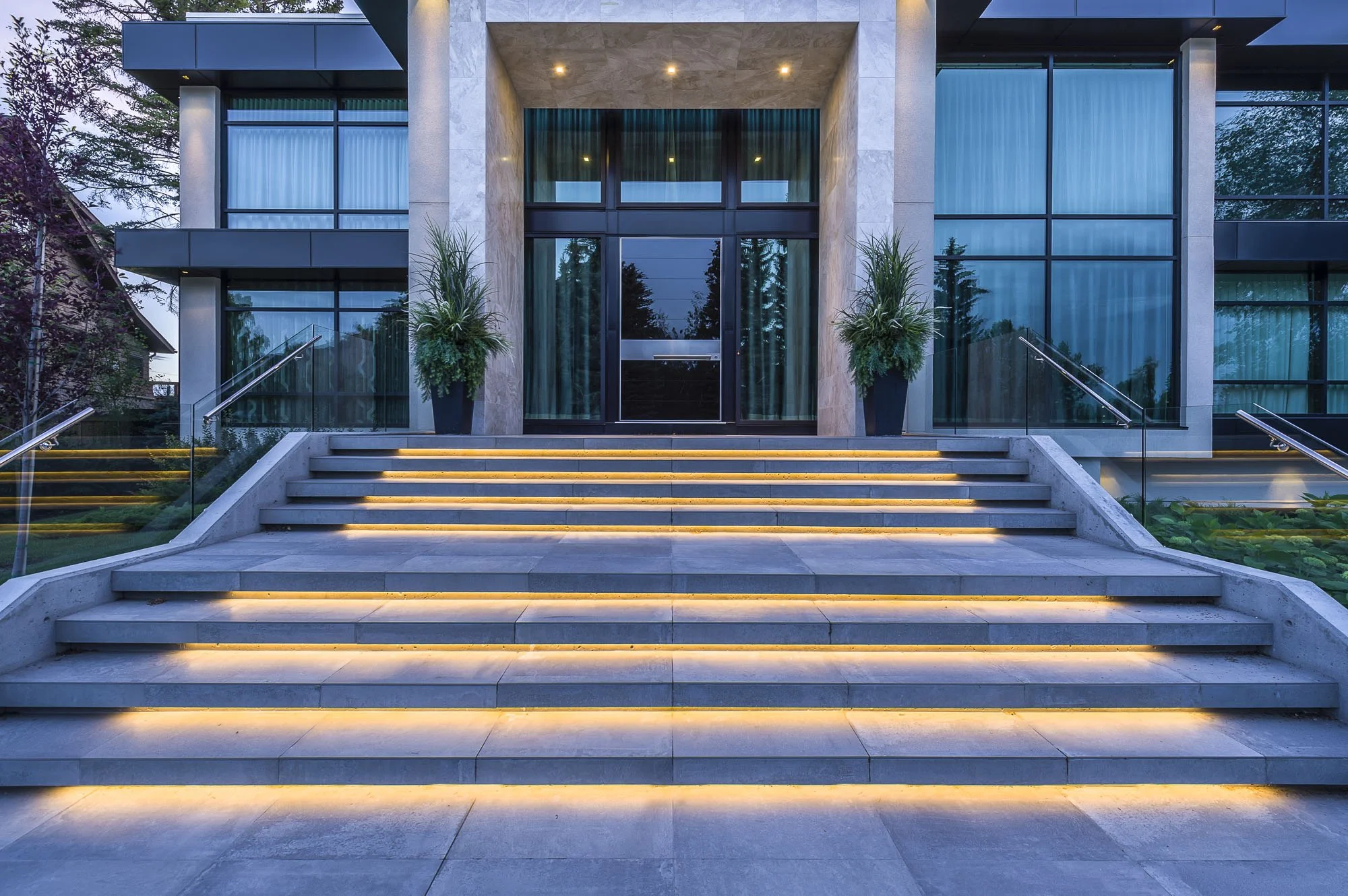 The entrance of a modern building with a set of wide stairs leading up to glass doors, flanked by large potted plants. The staircase is illuminated with warm lighting.