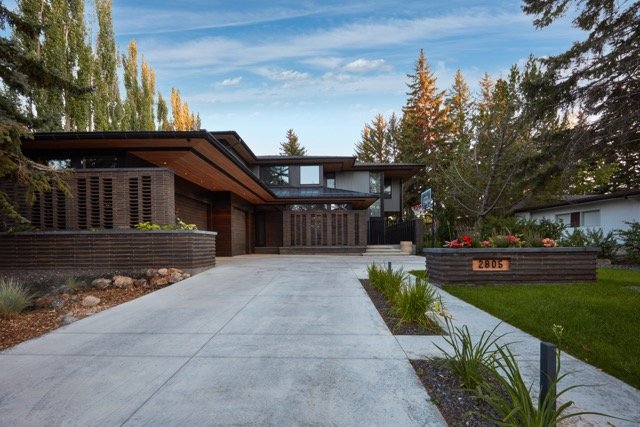Modern house with flat roof, large windows, and driveway surrounded by trees and landscaped yard.