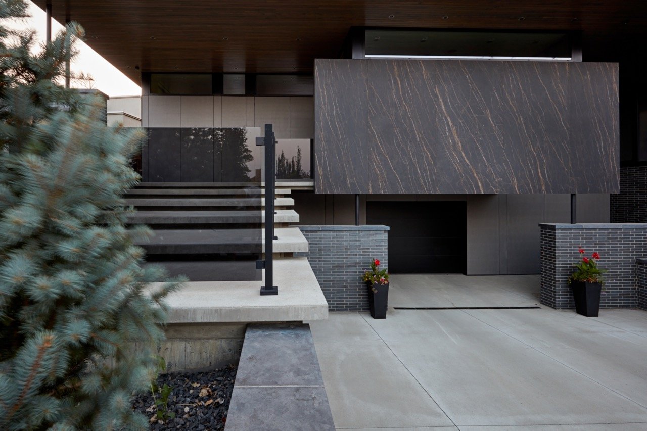 Modern house exterior with concrete steps and black railing, large dark marble facade, two brick planters with flowers, and a driveway.
