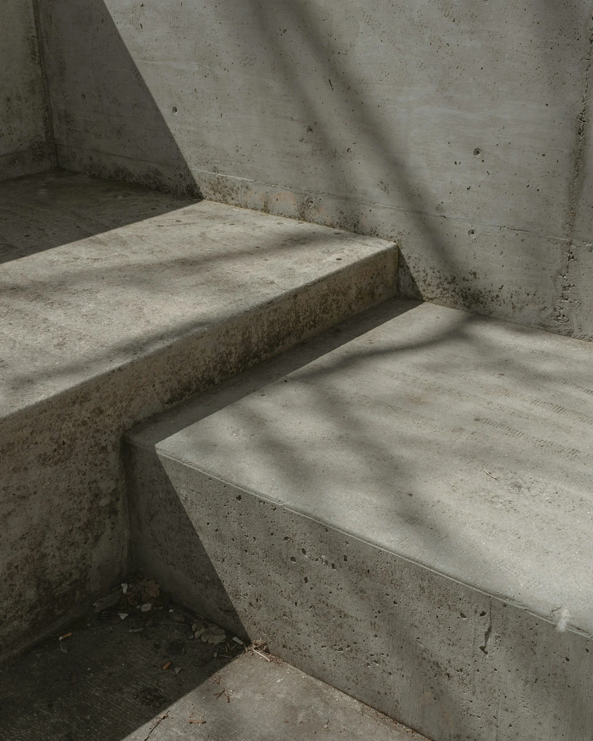 Concrete stairs and a concrete wall with some dirt, debris, and small holes, illuminated by natural sunlight creating shadows.