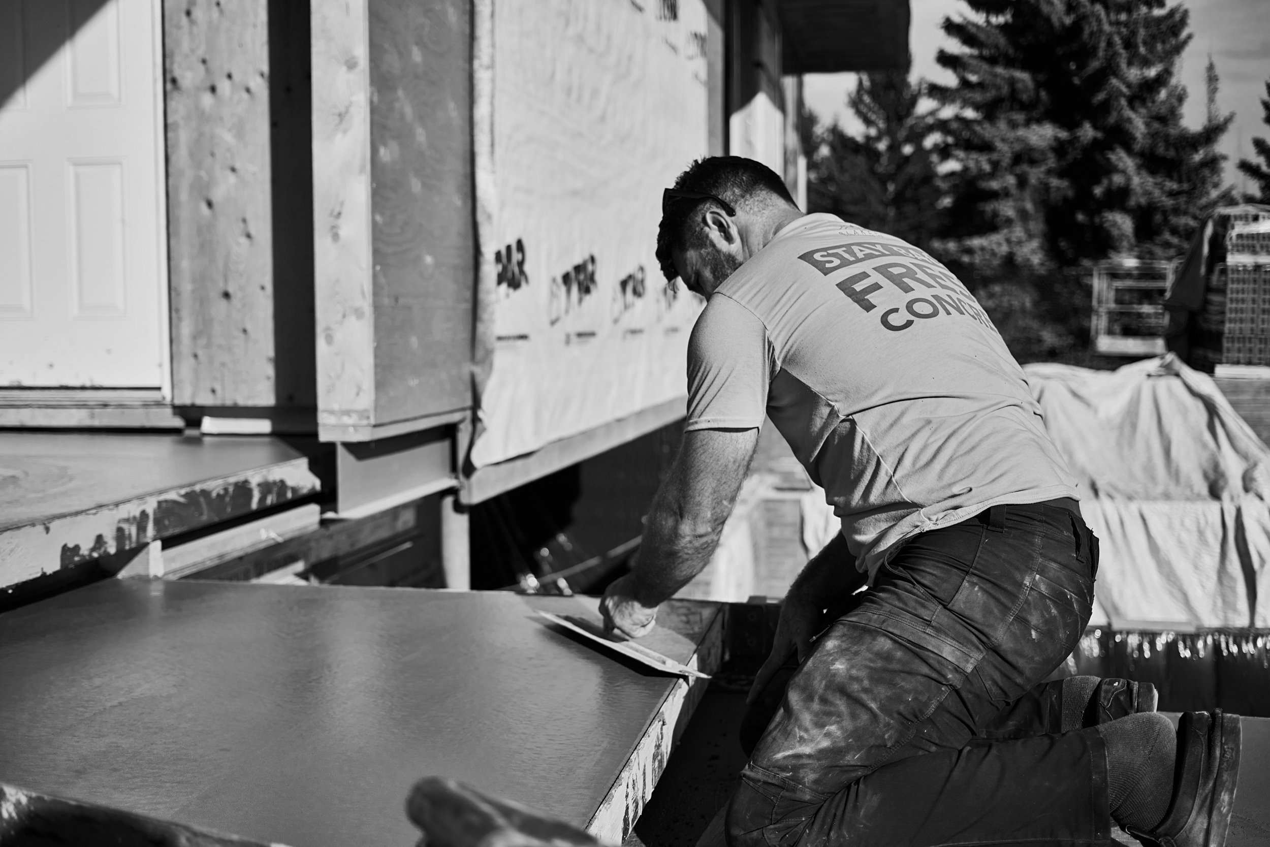 A man working on a construction site, installing a wooden panel on a building exterior. He is kneeling on the ground, wearing a t-shirt with the words 'Stay Focused' on the back, and appears to be using a tool to fit the panel.