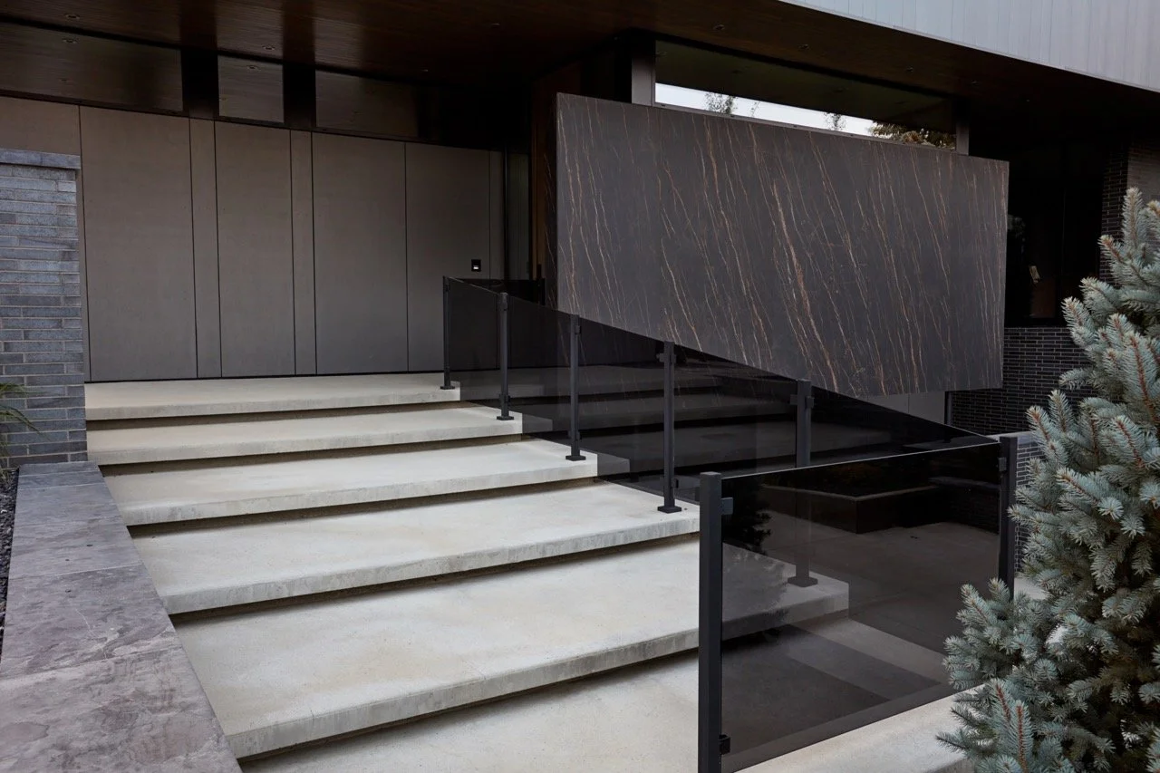 Modern house entrance with beige concrete stairs, black metal railing, dark wooden wall panel, and large black marble wall with orange veining, surrounded by evergreen trees.
