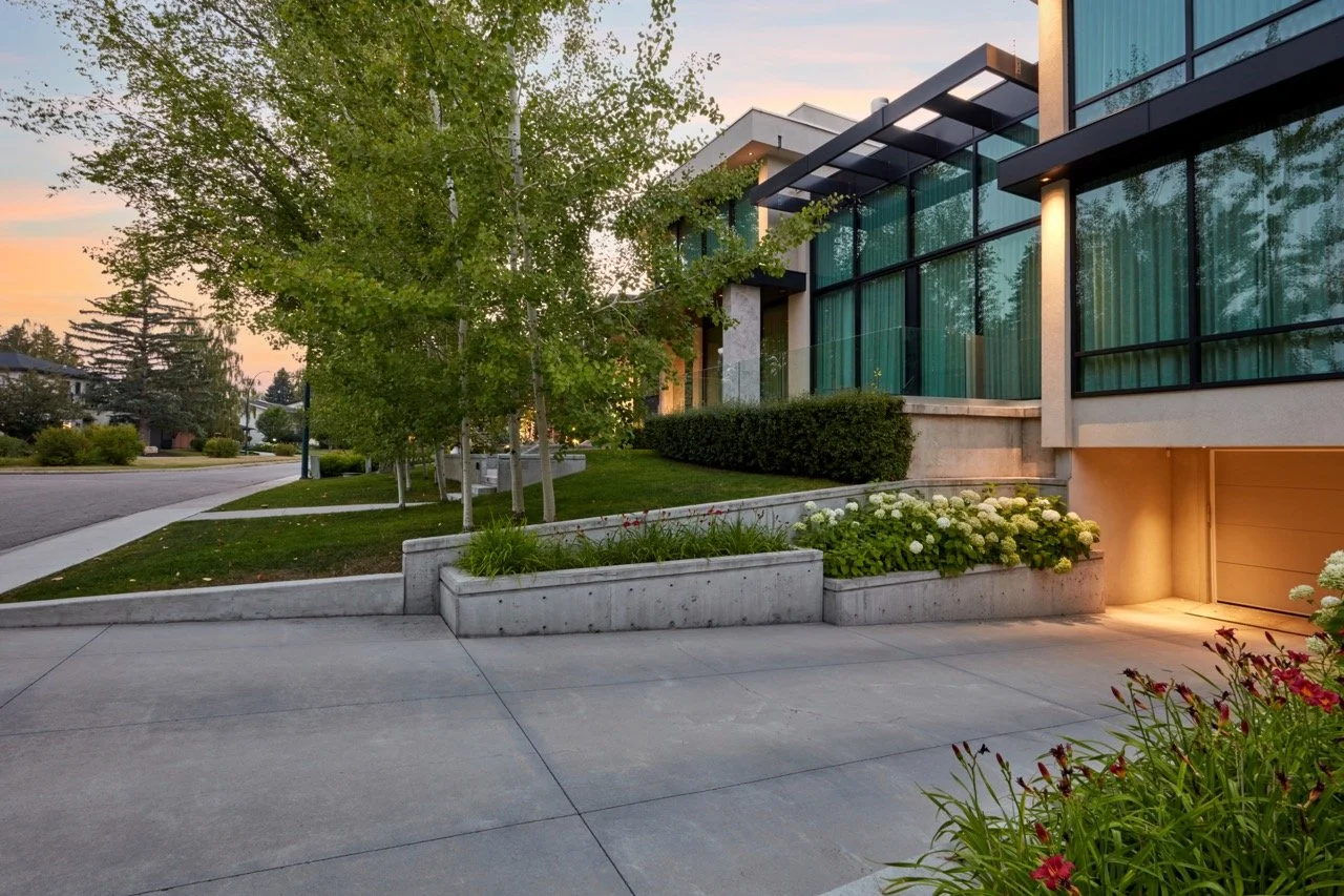 Modern house with large glass windows, surrounded by greenery and landscaped yard at sunset.