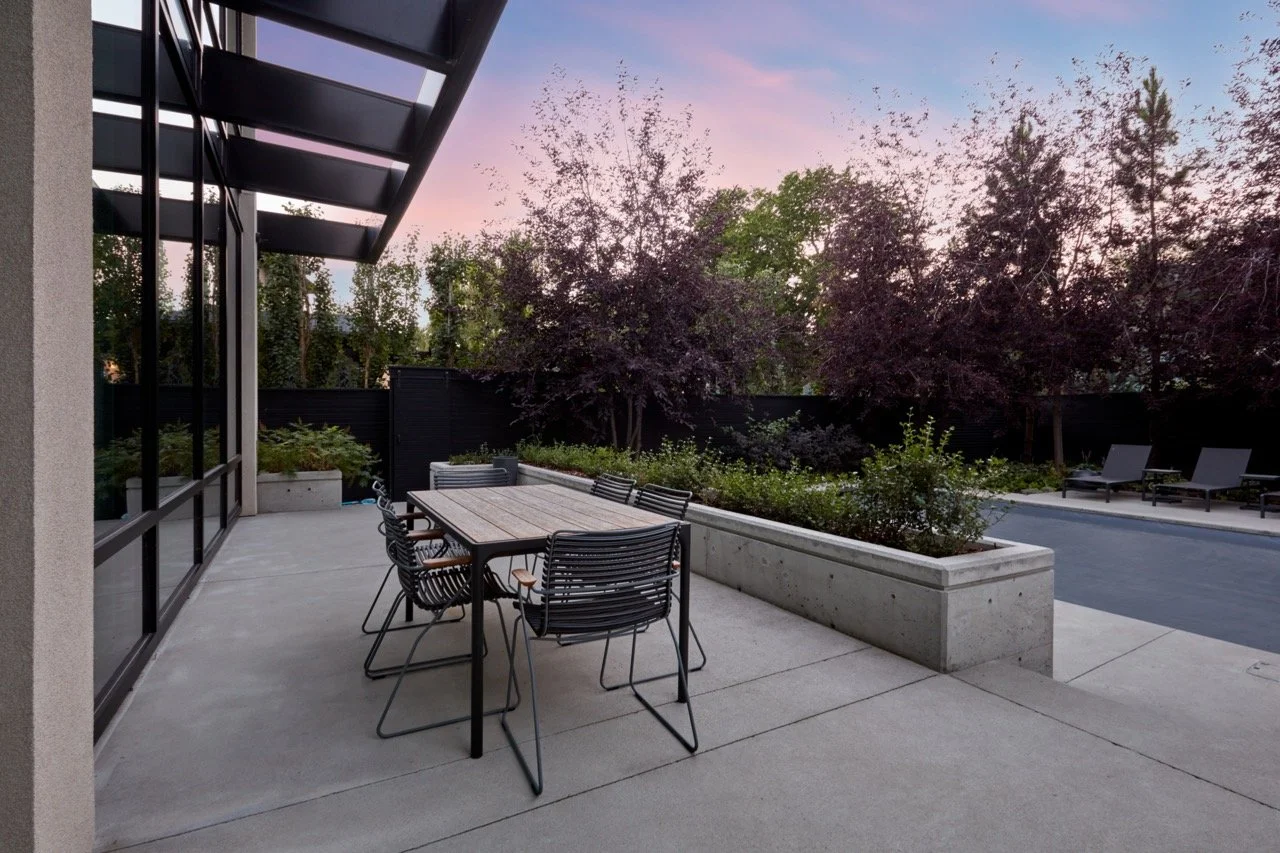 Modern outdoor patio with a wooden dining table and black metal chairs, surrounded by lush trees and plants at dusk.