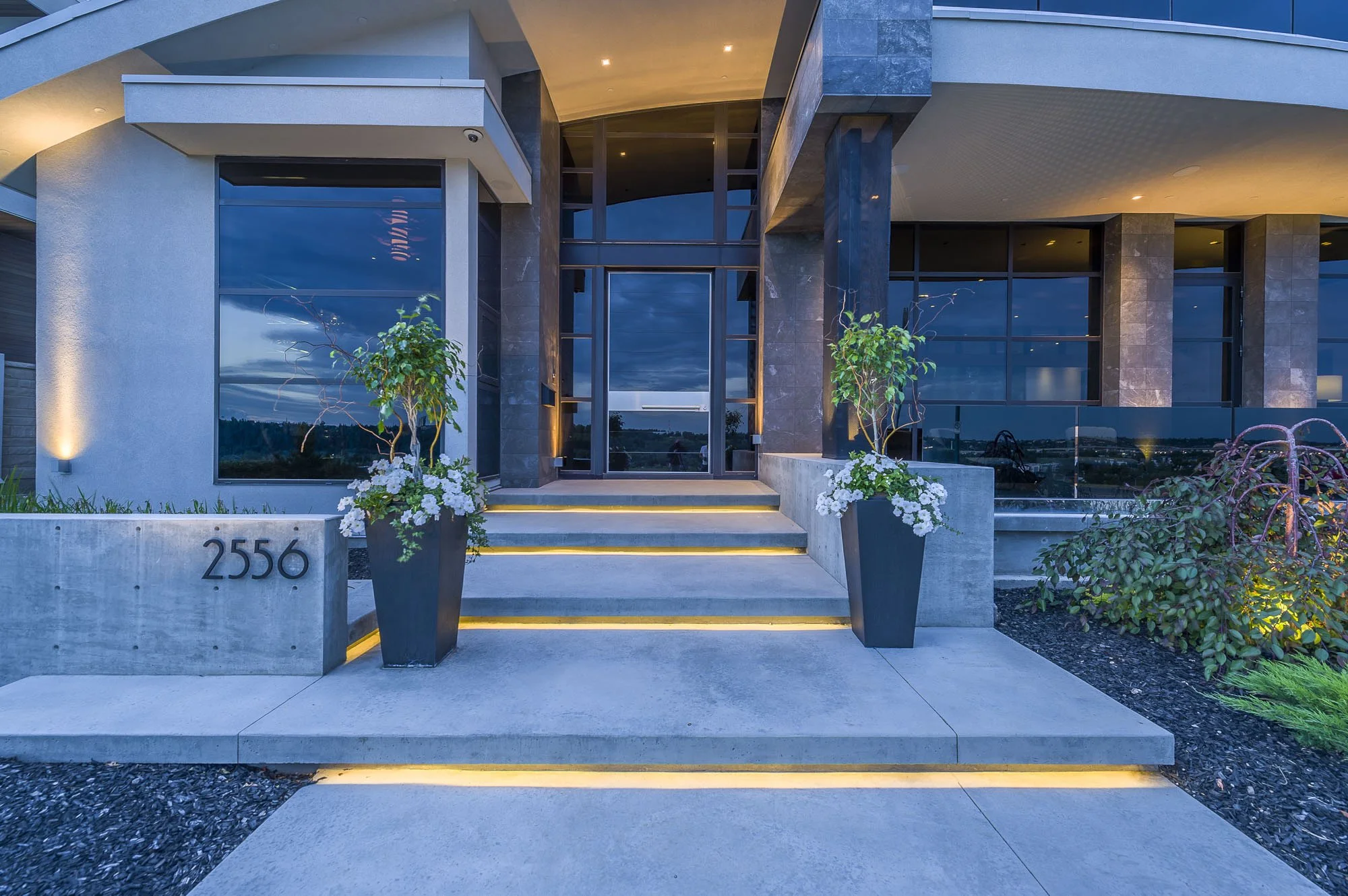 Modern house with illuminated steps, potted plants, large glass windows, and a covered entrance area at dusk.