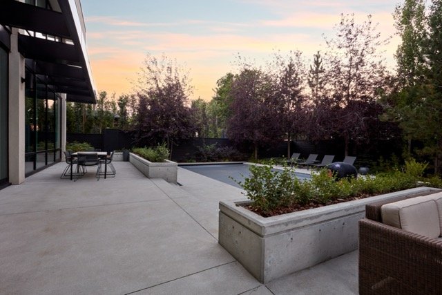 Modern patio with concrete planters, outdoor seating, and pool; surrounded by trees and soft evening light.