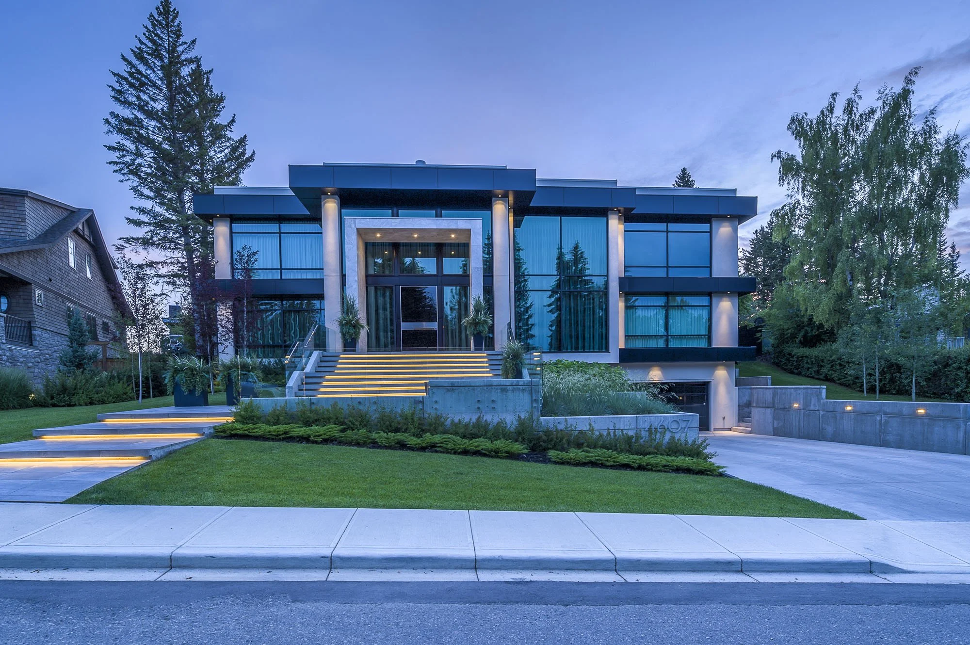 Modern two-story house with large glass windows, front steps with built-in lighting, surrounded by trees and greenery during dusk.