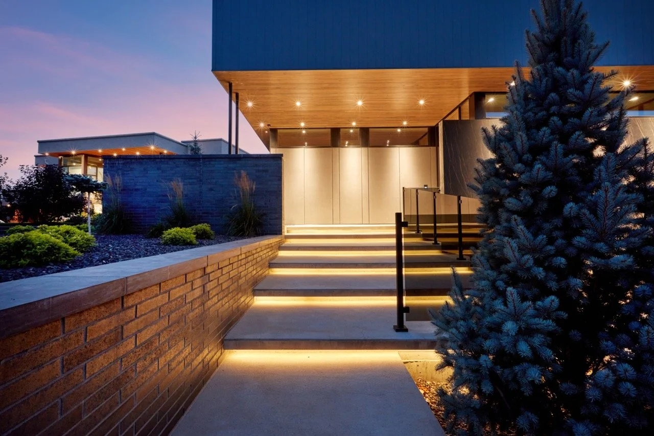 Modern house entrance illuminated at dusk with steps, brick wall, and surrounding landscaping including a large pine tree.