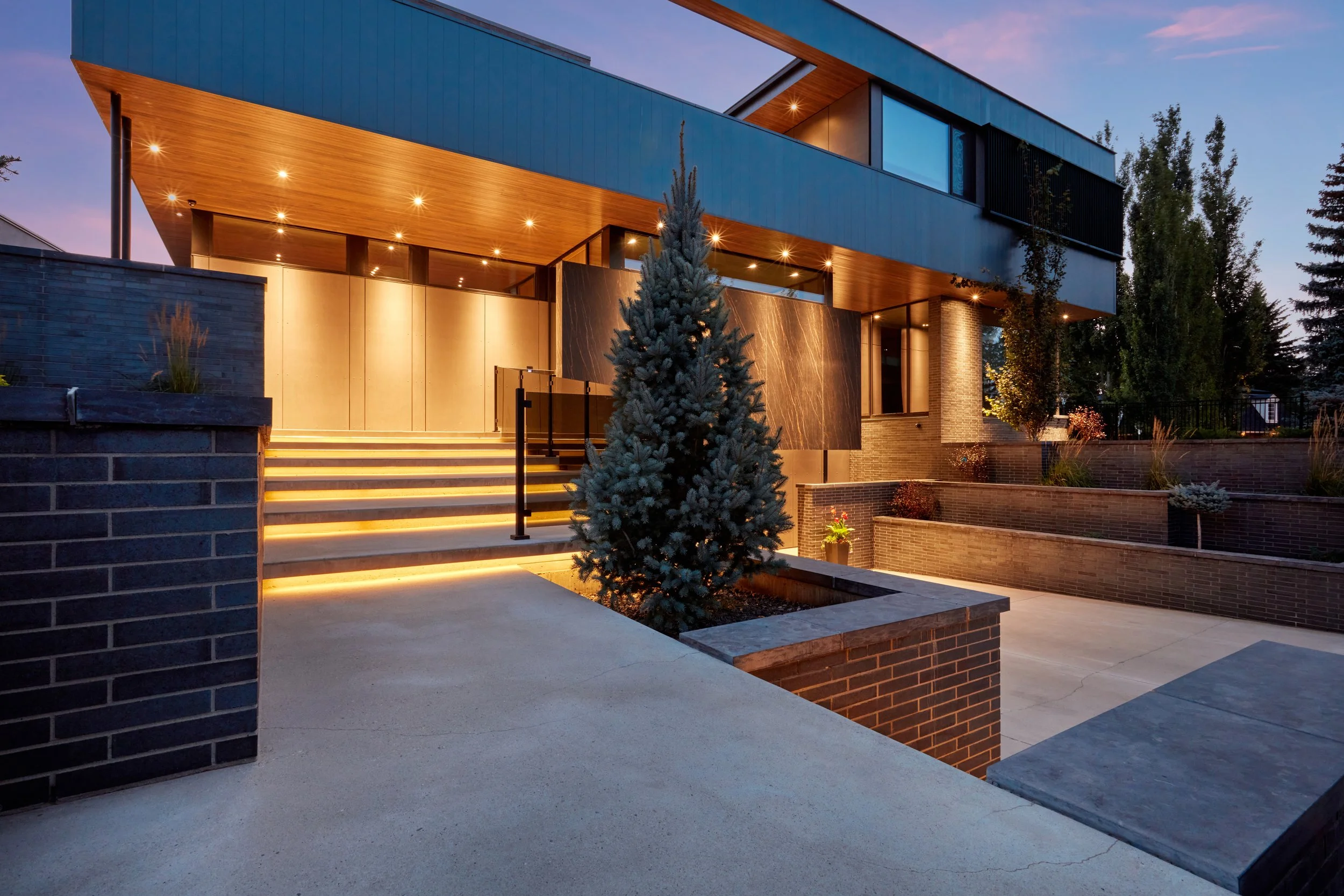Modern two-story house with large windows, brick and wood exterior, surrounded by trees, illuminated by exterior lighting at dusk.