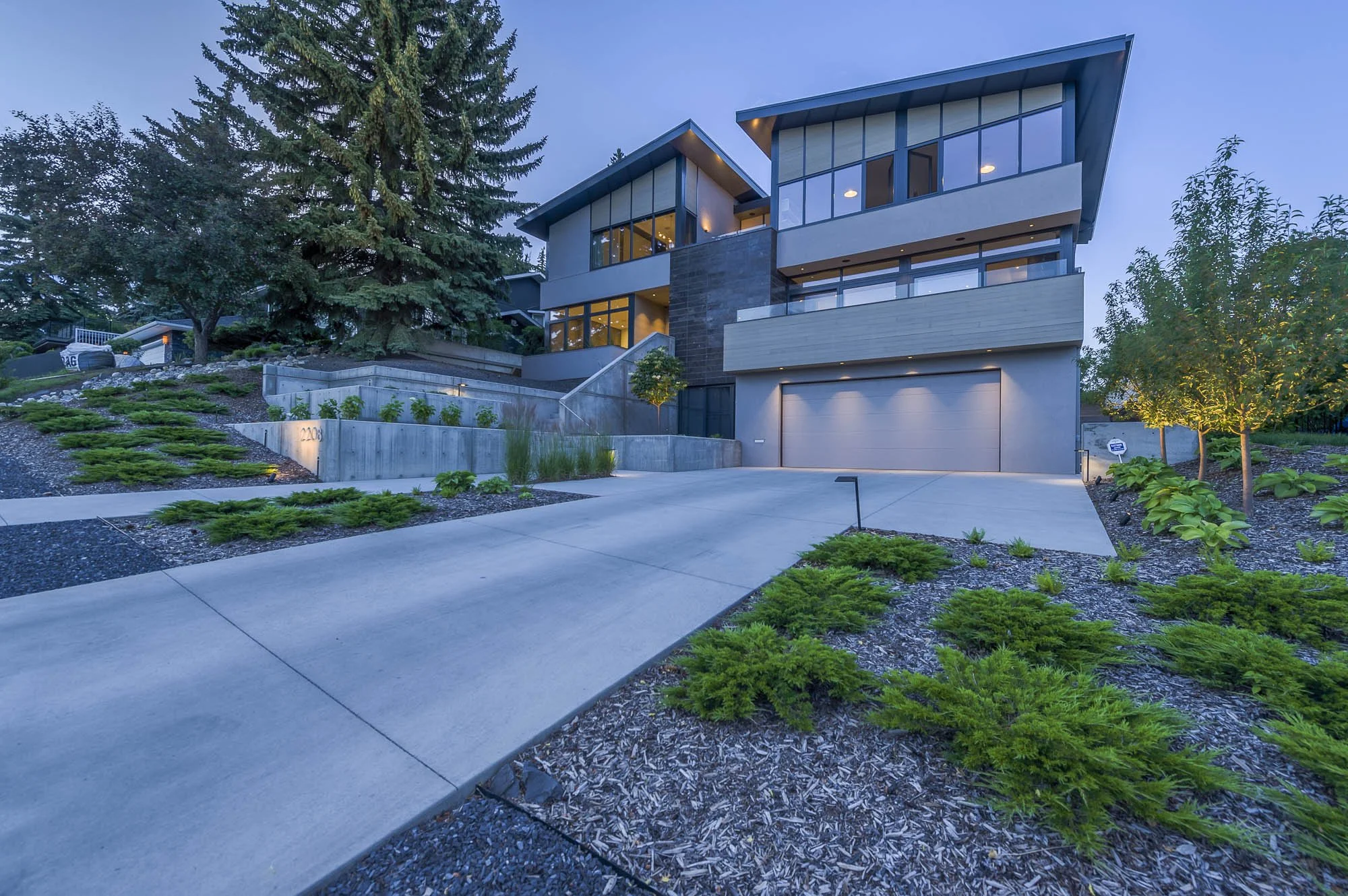 Modern multi-story house with large glass windows, surrounded by trees and landscaped yard at dusk.