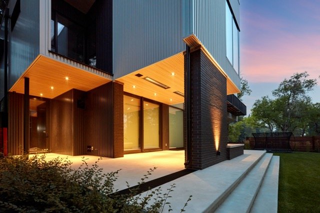 Modern house exterior with large windows, brick and wood siding, recessed lighting, and a spacious patio at dusk.