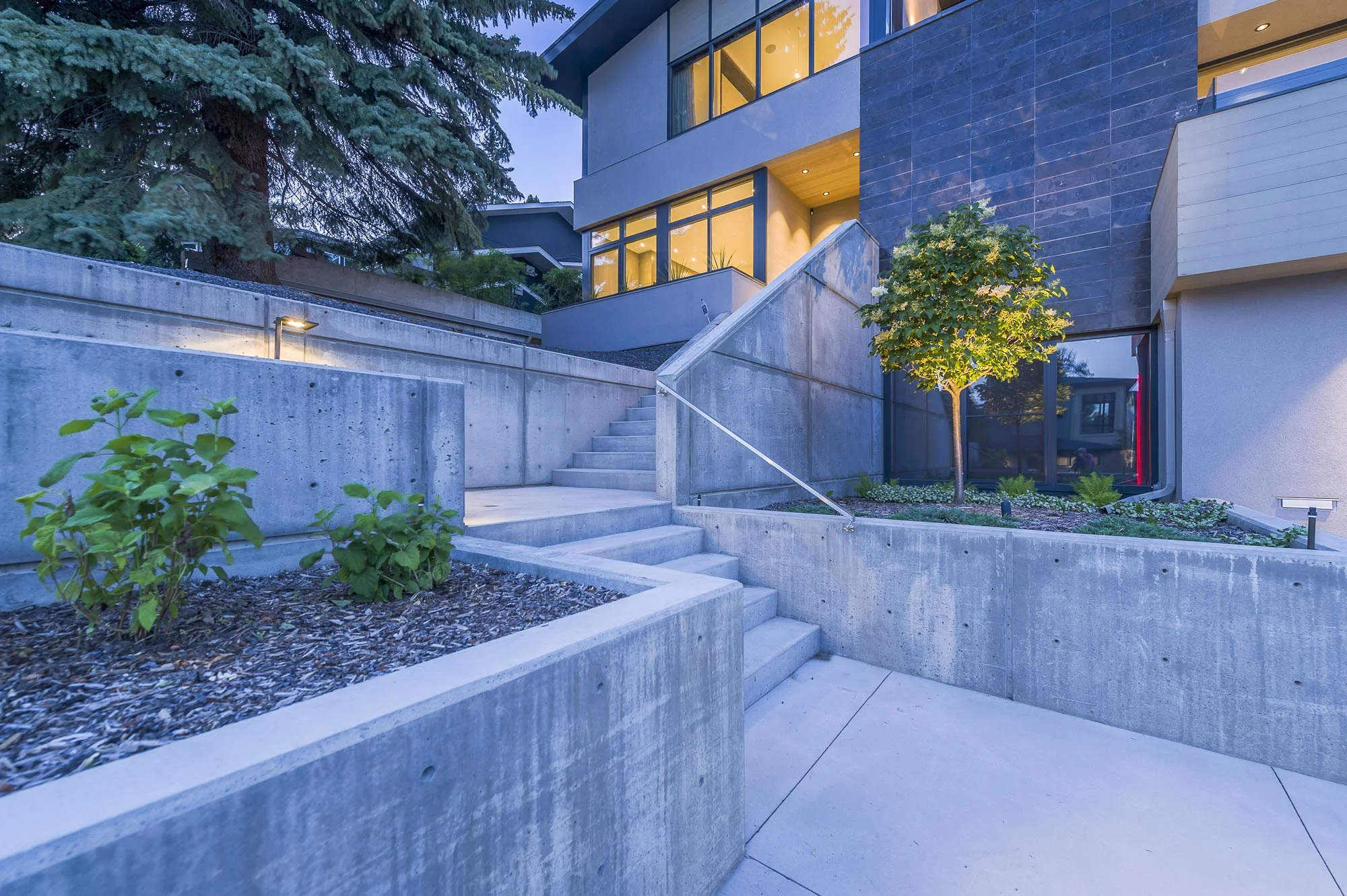 Modern residential building with large windows, concrete stairs, and landscaped greenery at dusk, illuminated by exterior lighting.