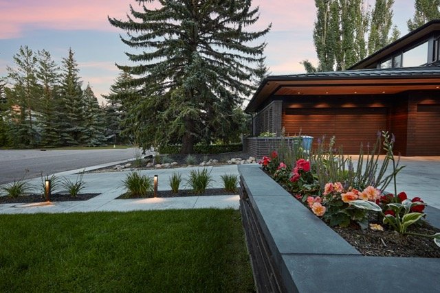 Modern house exterior with a landscaped front yard, featuring a lawn, flower beds with red and orange flowers, and path lights. A wooden garage is visible, surrounded by tall evergreen trees.