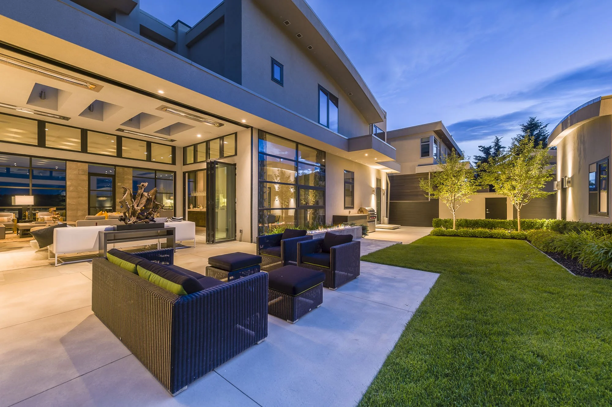 Modern multi-story house with large glass windows and outdoor seating area, illuminated at dusk. Green lawn with small trees surrounds the porch area.