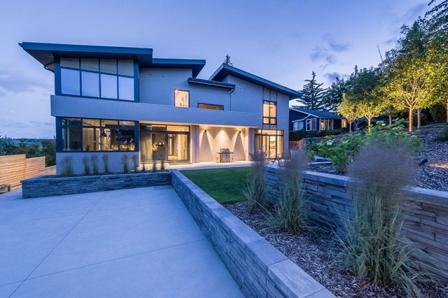 Modern two-story house with large windows and angular design, surrounded by landscaped gardens and trees. The house features a well-lit entrance and concrete driveway, with decorative grasses lining the pathway.