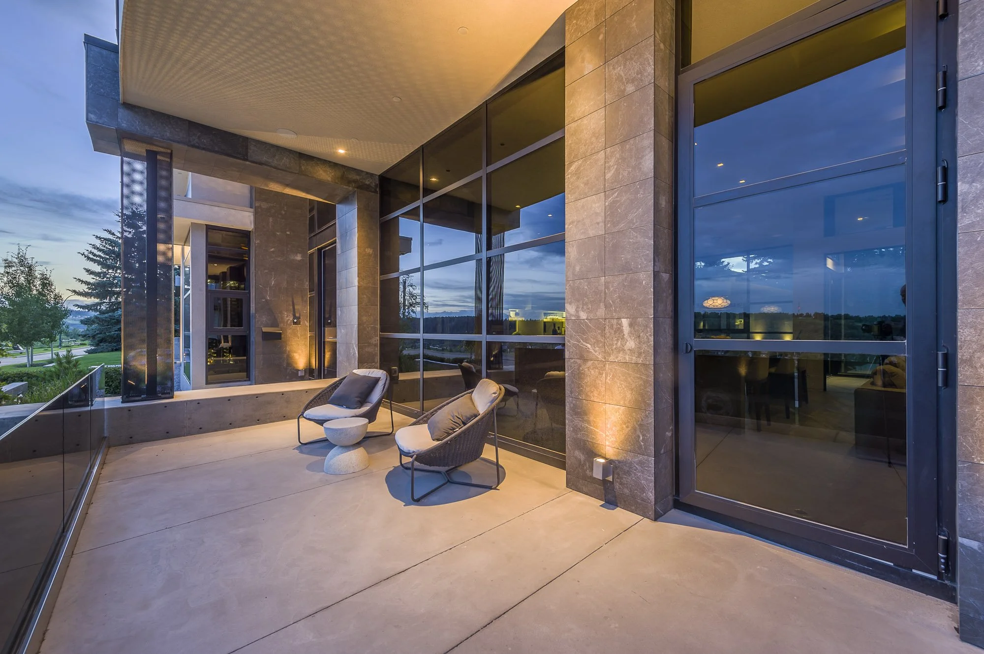 Modern balcony with lounge chairs, a small table, large glass windows, and warm exterior lighting at dusk.