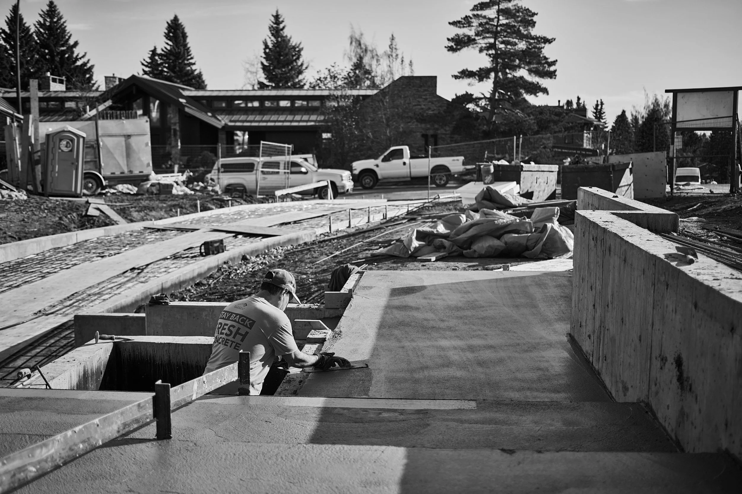Construction workers laying concrete on a building foundation outside, with parked trucks and trees in the background.