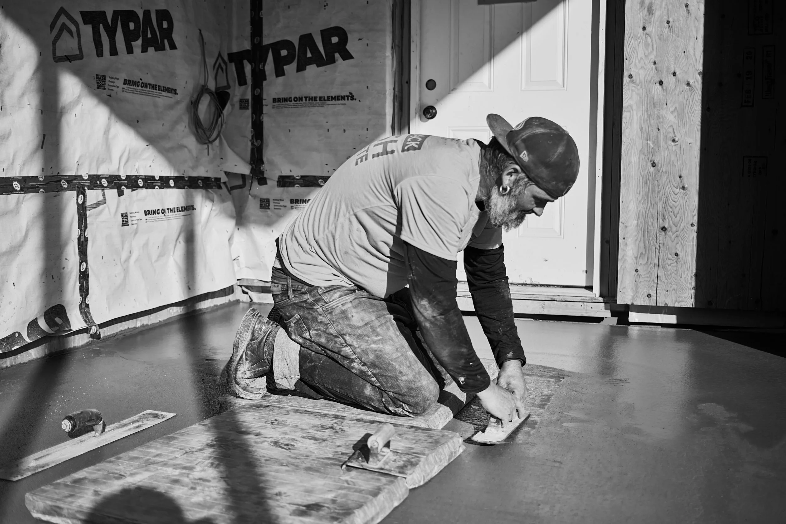 A man kneeling on the floor working on a construction or renovation project indoors. He is wearing a cap, earrings, a t-shirt, and jeans. The background shows unfinished wall covered with Typar weather-resistant barrier, some tools, and a door.
