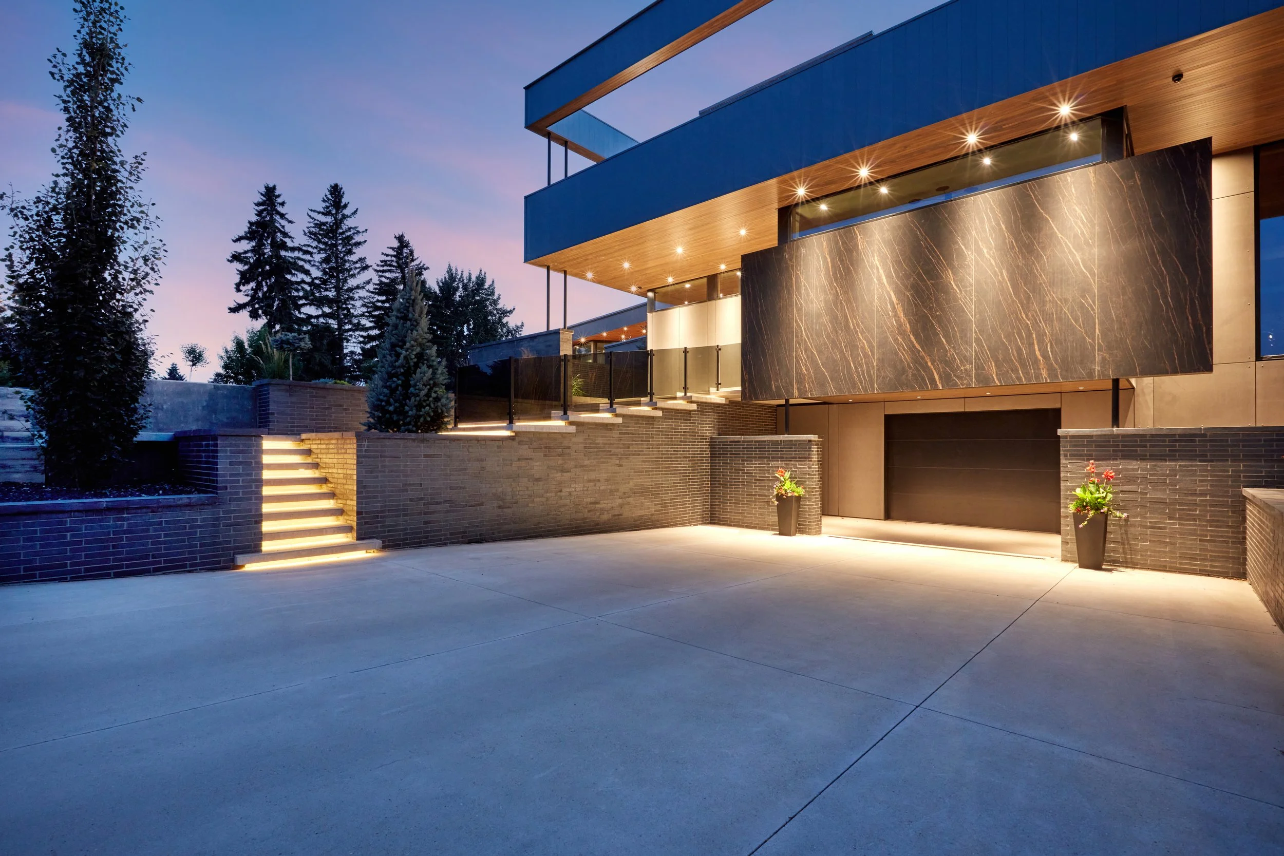 Modern house with exterior lighting, large marble wall, brick stairs, and surrounded by trees during dusk.