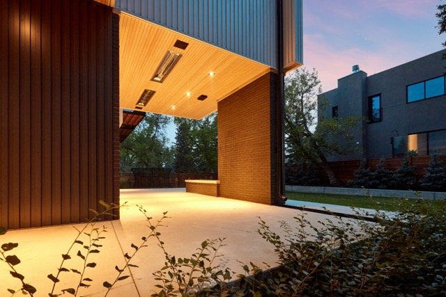 Modern architectural design of a house with an open, covered patio area, featuring wooden ceiling panels and recessed lighting. The exterior walls have vertical cladding, and the scene includes surrounding trees and a neighboring house against a dusk