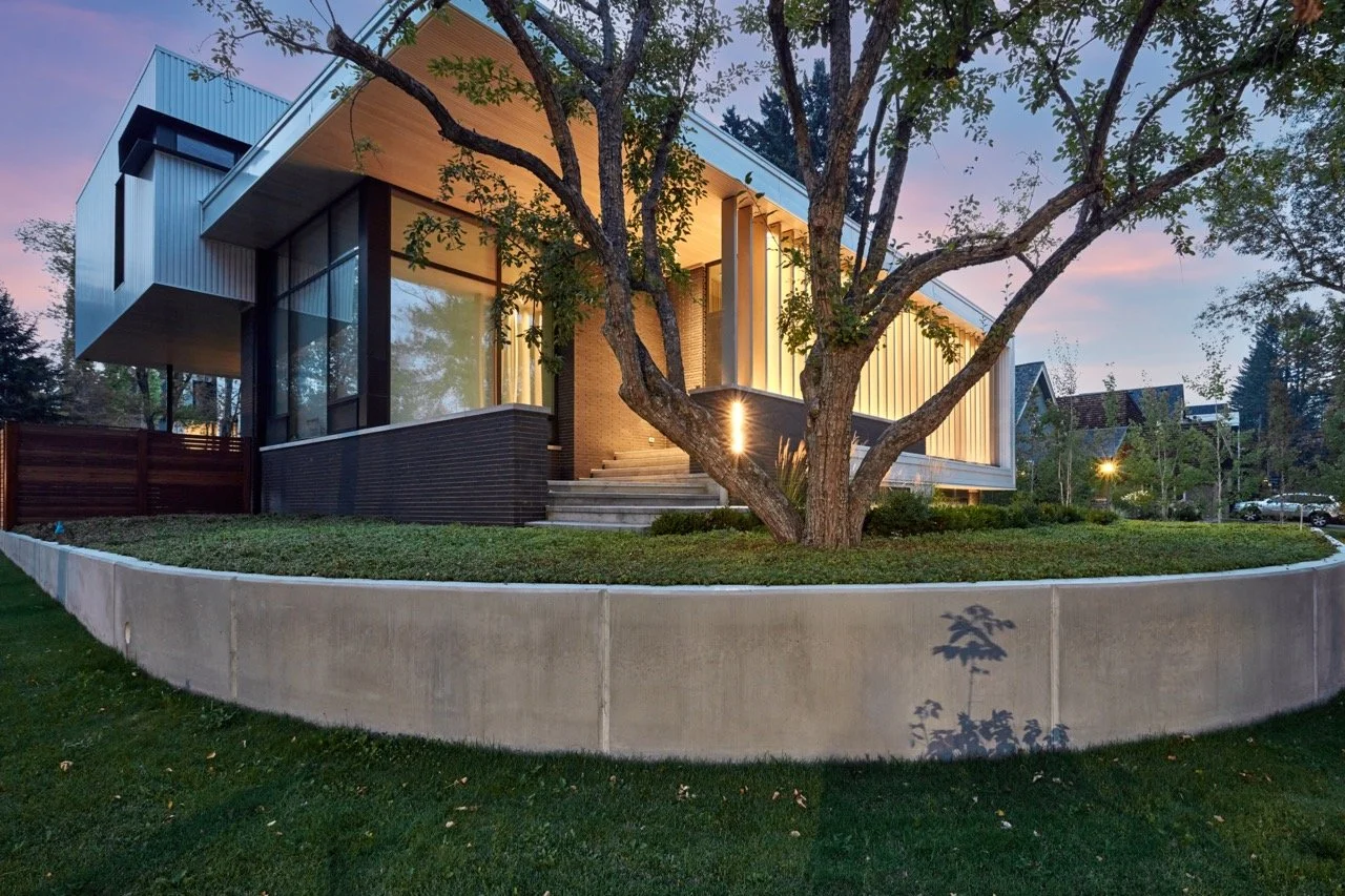 Modern house with large glass windows illuminated at dusk, surrounding landscaped yard with a prominent large tree and a concrete retaining wall.