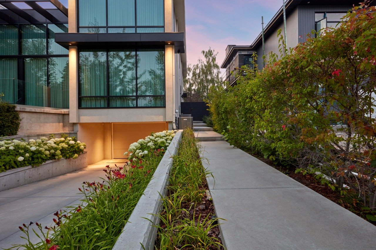 Modern residential building with large glass windows and a concrete walkway, surrounded by landscaped bushes and flowers during sunset.