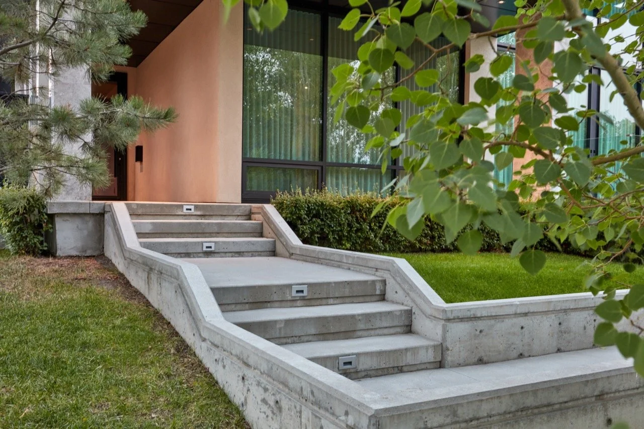 Concrete outdoor staircase leading to a modern building with large glass windows, surrounded by green grass, bushes, and trees.