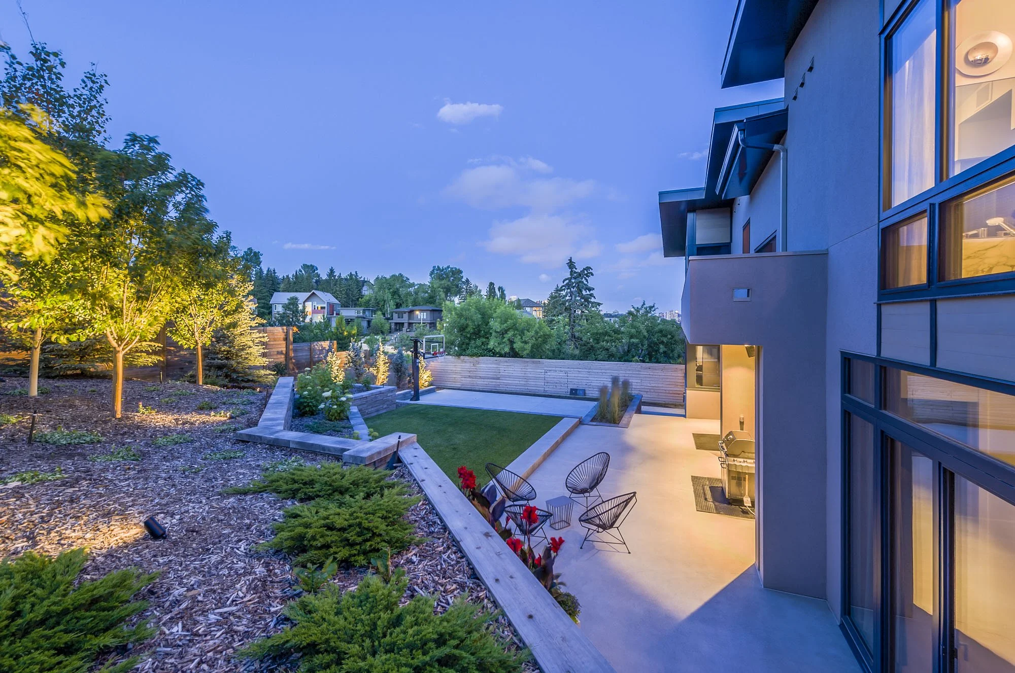 Night view of a modern house's backyard with illuminated outdoor seating area, trees, and neighboring houses in the background.