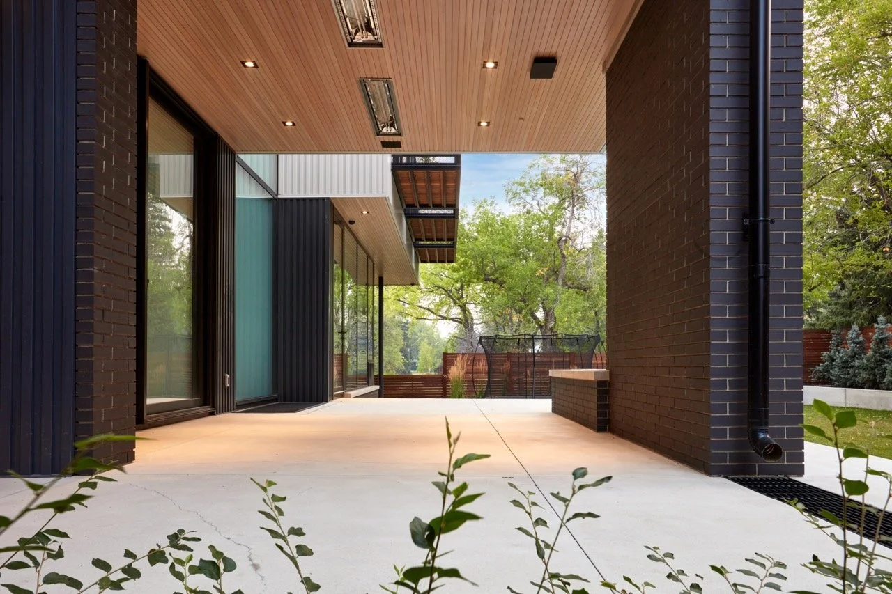 Modern house exterior with a covered patio, black brick walls, large glass sliding doors, wooden ceiling, outdoor lighting, and greenery in the background.