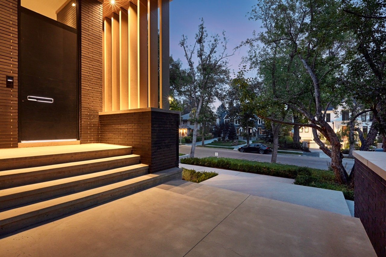 Front steps and porch of a modern house at dusk, with brick walls, wooden accents, and trees in the yard.
