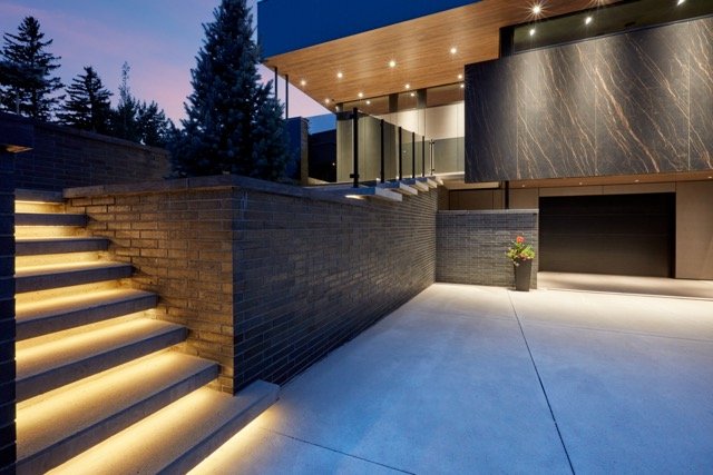 Modern house exterior with illuminated steps, dark brick walls, large windows, and a sleek garage door at dusk.