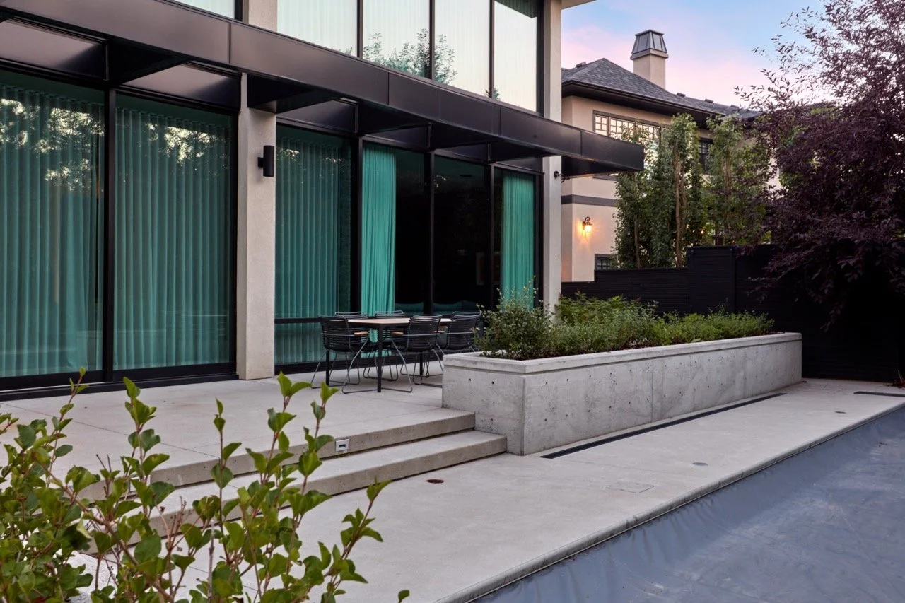 Modern house with large glass sliding doors, outdoor patio with black metal chairs and table, concrete steps, garden with shrubs and trees, and a privacy fence, during dusk.
