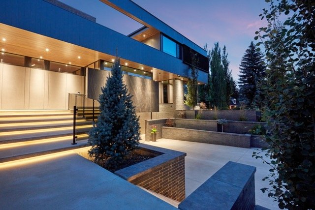 Modern house exterior at dusk with minimalist design, decorative stairs, ambient lighting, and landscaped trees and plants.