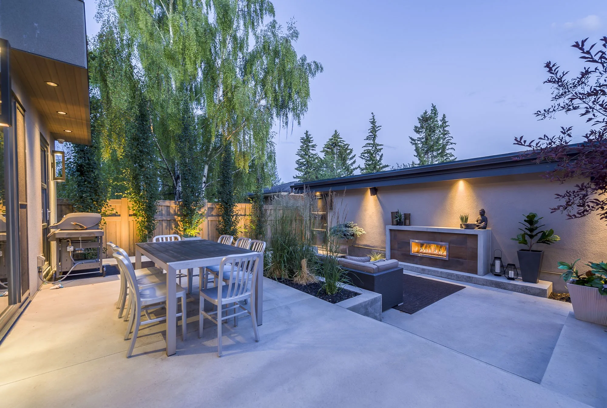 A backyard outdoor patio at dusk with a dining table and chairs, an outdoor fireplace with decorative items, and trees in the background.