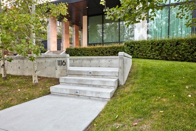 Concrete steps leading to a modern building with large glass windows, surrounded by trees and manicured hedges. The number 1116 is displayed on a concrete wall near the steps.