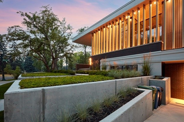 Modern building with large windows and wooden accents, surrounded by landscaped gardens and trees, during twilight.