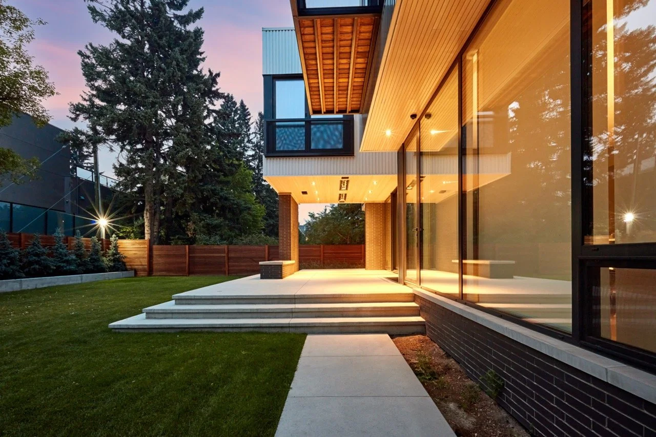 Modern house with large glass windows, wooden accents, and a spacious backyard with steps leading to a patio at dusk.